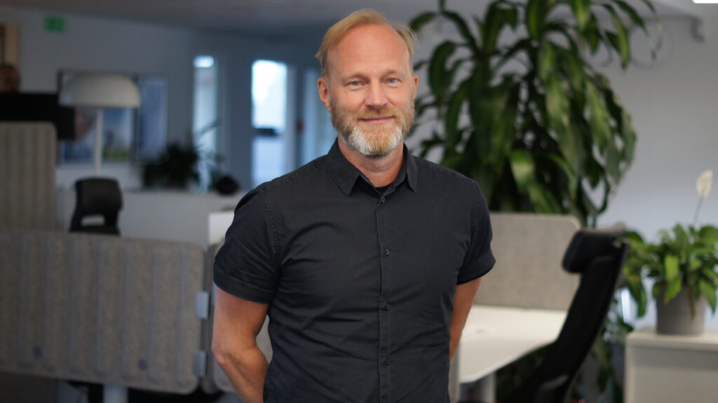 John Carlsson som är Pyramidkonsult på Wiig Data står i en modern kontorsmiljö med gröna växter och skrivbord i bakgrunden.